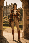 Luxury editorial image of a woman in a fitted brown tartan blazer walking through a historic town, styled in an elevated autumn outfit. The blazer is sharply cut, smooth and well structured, paired with complementary separates for a sophisticated fall look inspired by heritage fashion and timeless city elegance.